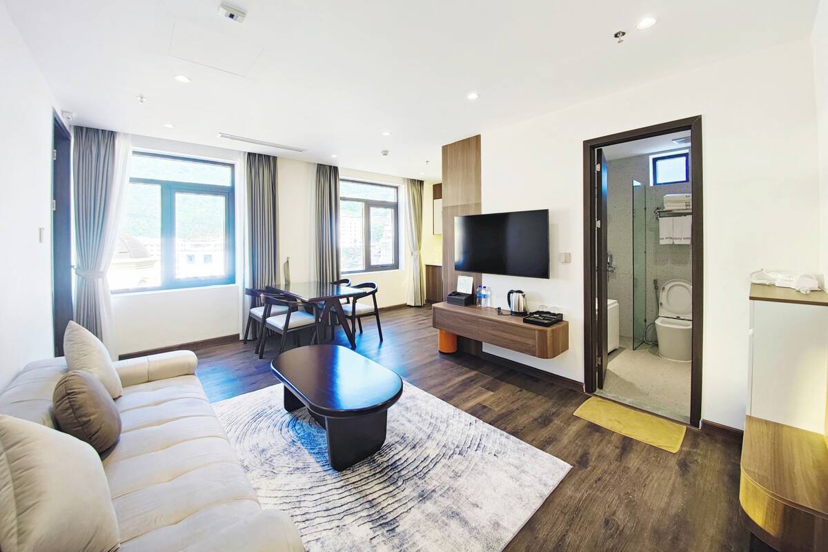 Une chambre d’hôtel moderne équipée d’un canapé, d’une table à manger avec des chaises, d’un téléviseur à écran plat, d’un petit bureau et d’une salle de bain visible à travers une porte en verre.