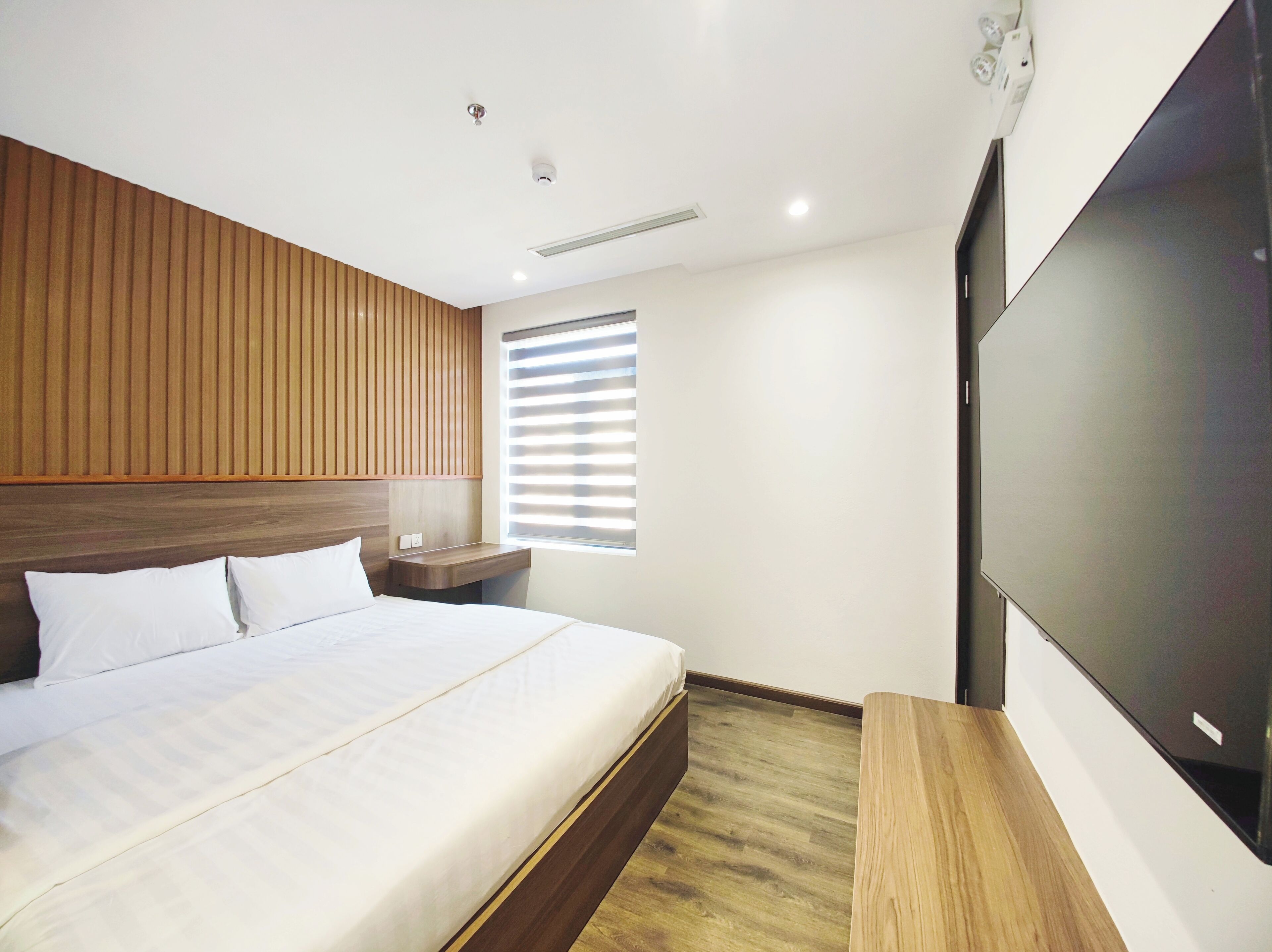 A modern hotel room with a large bed, wooden headboard, a flat-screen TV, and a window with blinds.