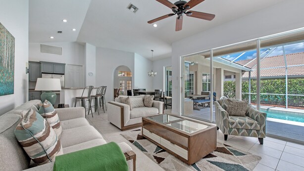 A spacious living room with a ceiling fan, a glass coffee table, and a view of a swimming pool through large sliding glass doors.