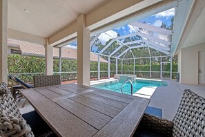 Ein überdachter Poolbereich im Freien mit einem Holztisch und Korbmöbeln, umgeben von einer Glaseinfassung.
