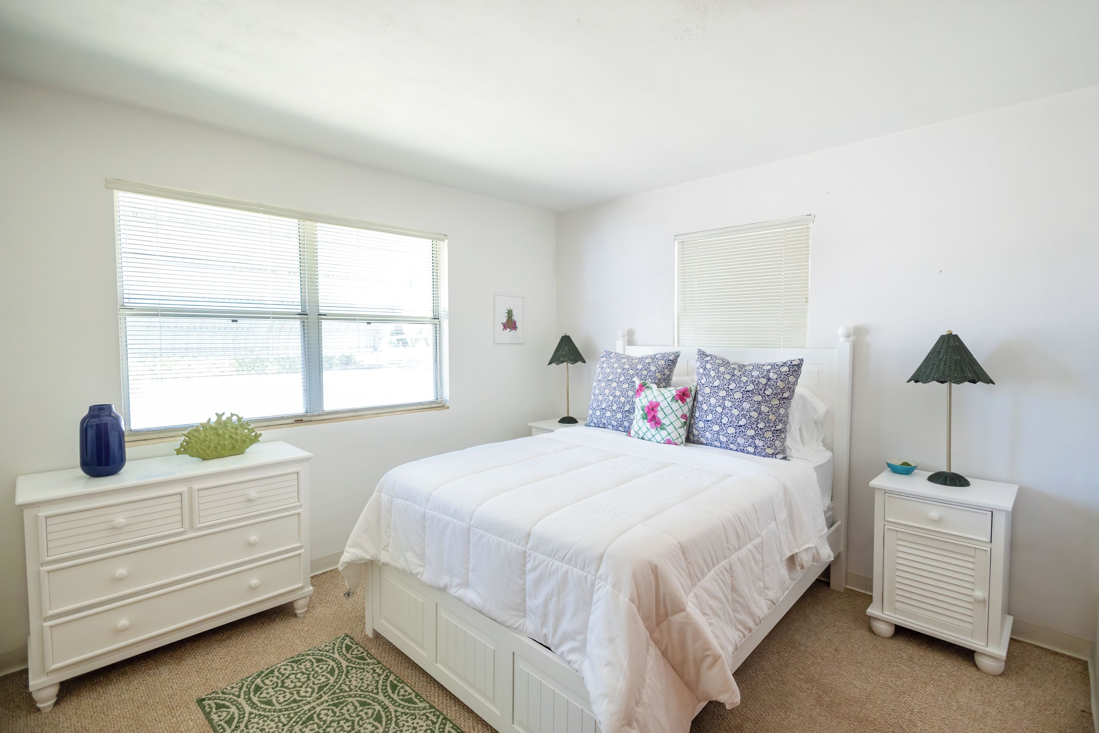 A bedroom with a white bed, two nightstands, a dresser, and a window with blinds.