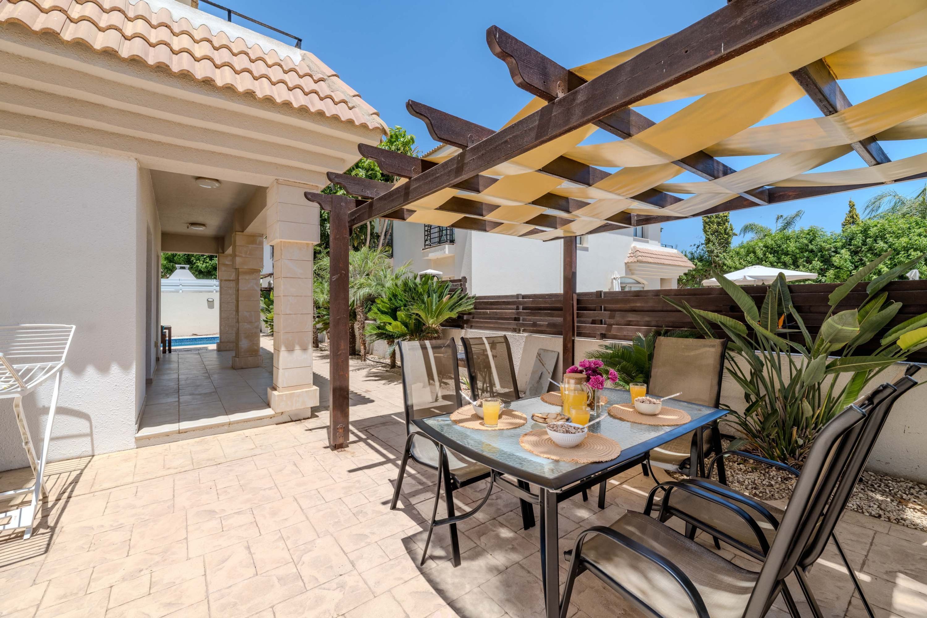 An outdoor patio with a table set for two, a pergola providing shade, and a view of a swimming pool in the background.
