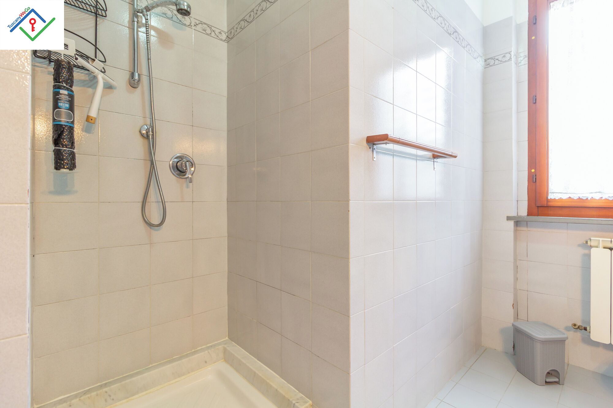 Un baño con paredes de azulejos blancos, una ducha con cabezal de ducha manual, un perchero y una ventana con marco de madera.