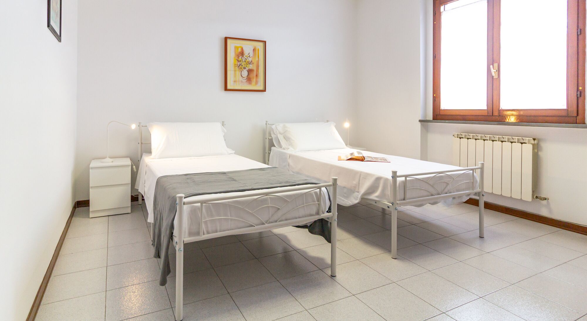 Una stanza con due letti, un piccolo comodino, un termosifone e una finestra con persiane in legno.
