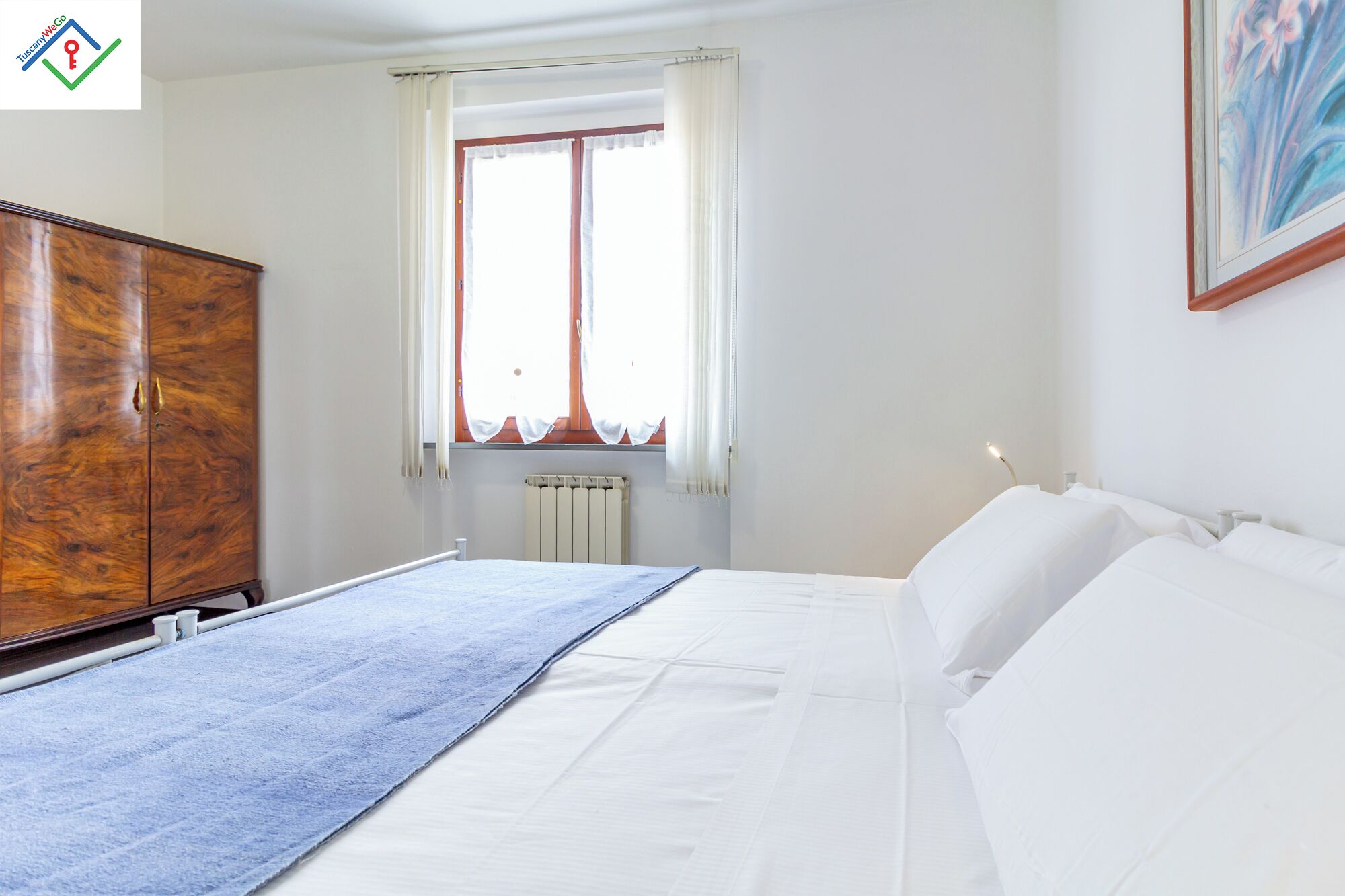 Un dormitorio con cama, armario de madera, radiador y ventana con cortinas.