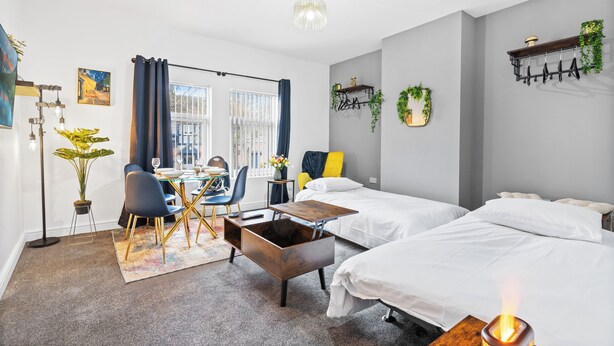 A modern hotel room with two beds, a dining area with a table and chairs, a potted plant, and a ceiling light.