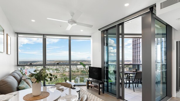 Una sala de estar moderna con comedor, ventanas amplias que ofrecen vistas de la ciudad, un ventilador de techo y un puerta de vidrio que da a un balcón exterior.