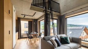 A modern living room with a sofa, dining table, and chairs. A wooden staircase leads to an upper level.