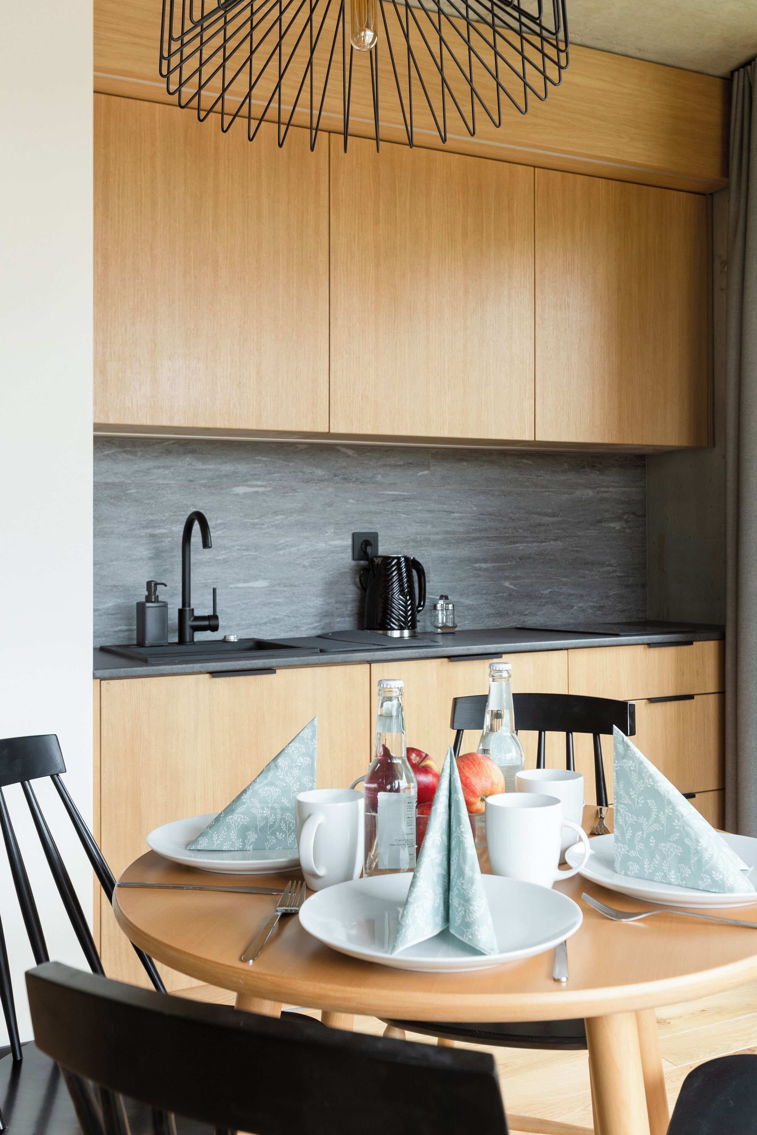Una cocina moderna con armarios de madera, un grifo negro y una encimera negra. Hay una mesa redonda con platos, tazas y una botella.