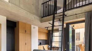A modern loft apartment with a loft bed, a grey sofa, a wooden coffee table, and a large window.