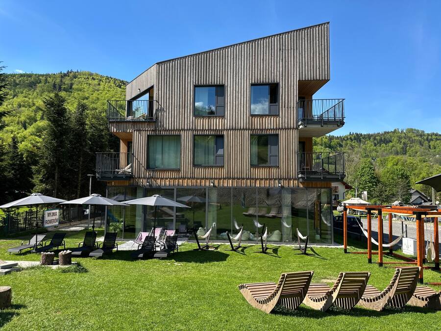 A modern multi-story building with a wooden facade, outdoor seating area with umbrellas, and a view of a forested hillside.
