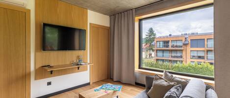 A modern living room with a flat-screen TV, wooden furniture, a large window with a view of buildings, and a cozy seating area.