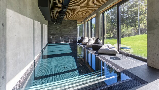 A modern indoor swimming pool with a glass floor, large windows, and a view of a green landscape.