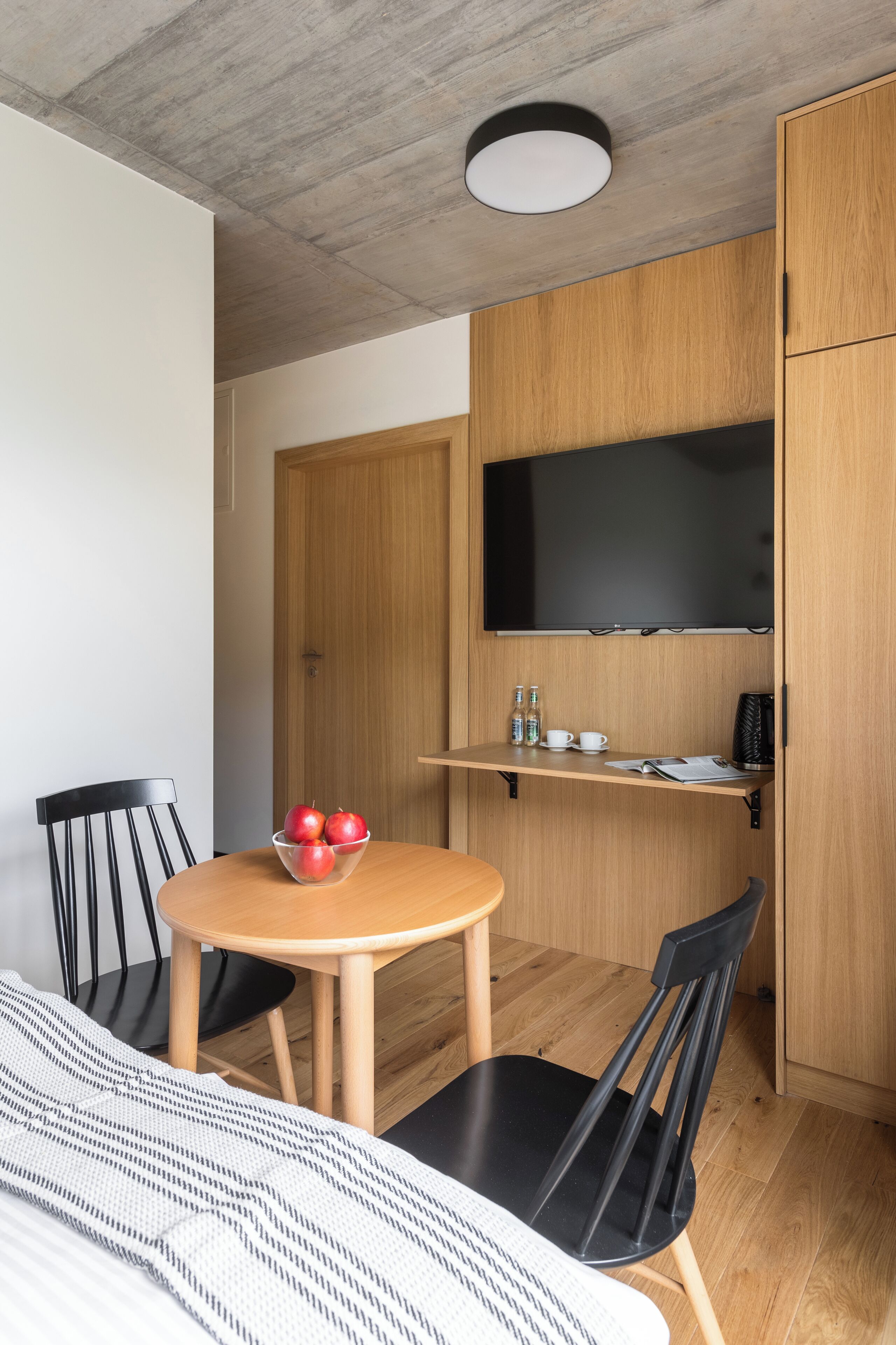 Habitación pequeña con una mesa redonda de madera, dos sillas negras, un cuenco de manzanas, un televisor de pantalla plana montado en un mueble de madera y una cama con una colcha a rayas.