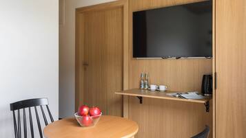 Habitación pequeña con una mesa redonda de madera, dos sillas negras, un cuenco de manzanas, un televisor de pantalla plana montado en un mueble de madera y una cama con una colcha a rayas.