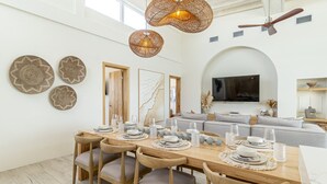Un comedor con una larga mesa de madera dispuesta para seis personas, rodeada de sillas. Hay lámparas colgantes tejidas sobre la mesa, un espejo grande en la pared y un sofá moderno al fondo.