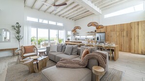Amplia sala de estar con un sofá grande, una mesa de centro y mesitas de noche de madera. Hay un comedor con una mesa y sillas de madera. La habitación cuenta con vigas de madera a la vista en el techo y ventanas grandes que proporcionan luz natural.