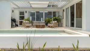 Un área de piscina moderna al aire libre con un porche cubierto, mobiliario y una parrilla.