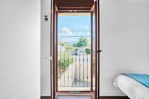 Una habitación con una puerta de vidrio corrediza que da a un balcón con baranda metálica, una cama con colchón blanco y ropa de cama azul, y una pequeña lámpara de pared.