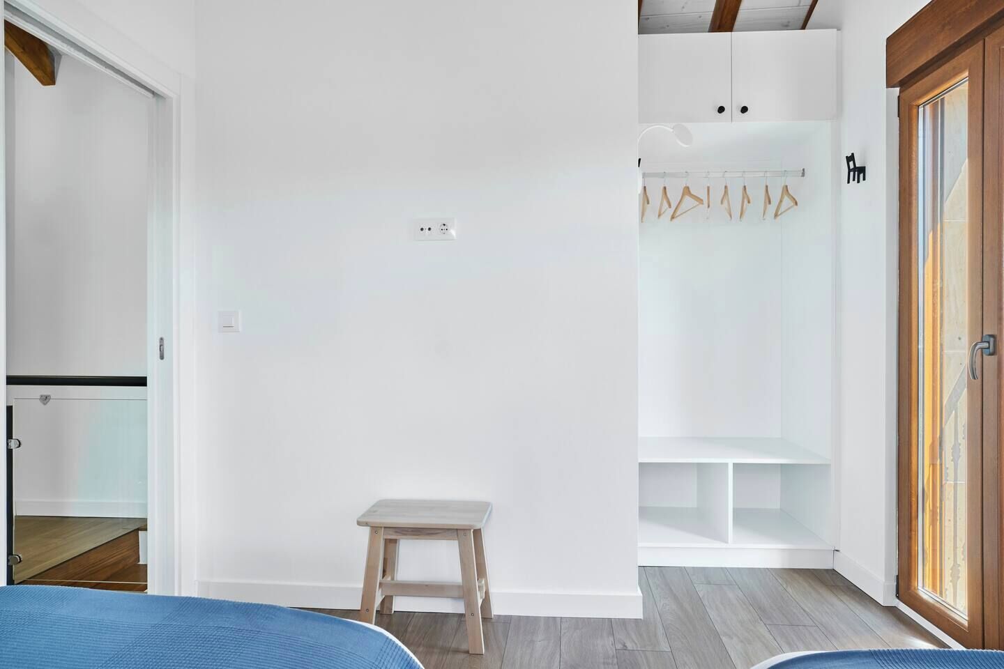 Un dormitorio con una cama, un taburete de madera, un armario y un closet con ropa colgada.