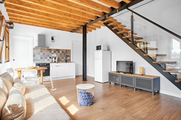 Eine moderne Küche mit Holzbalkendecke, einem Essbereich mit Tisch und Stühlen, einer Couch und einer Treppe mit Glastrennwand.