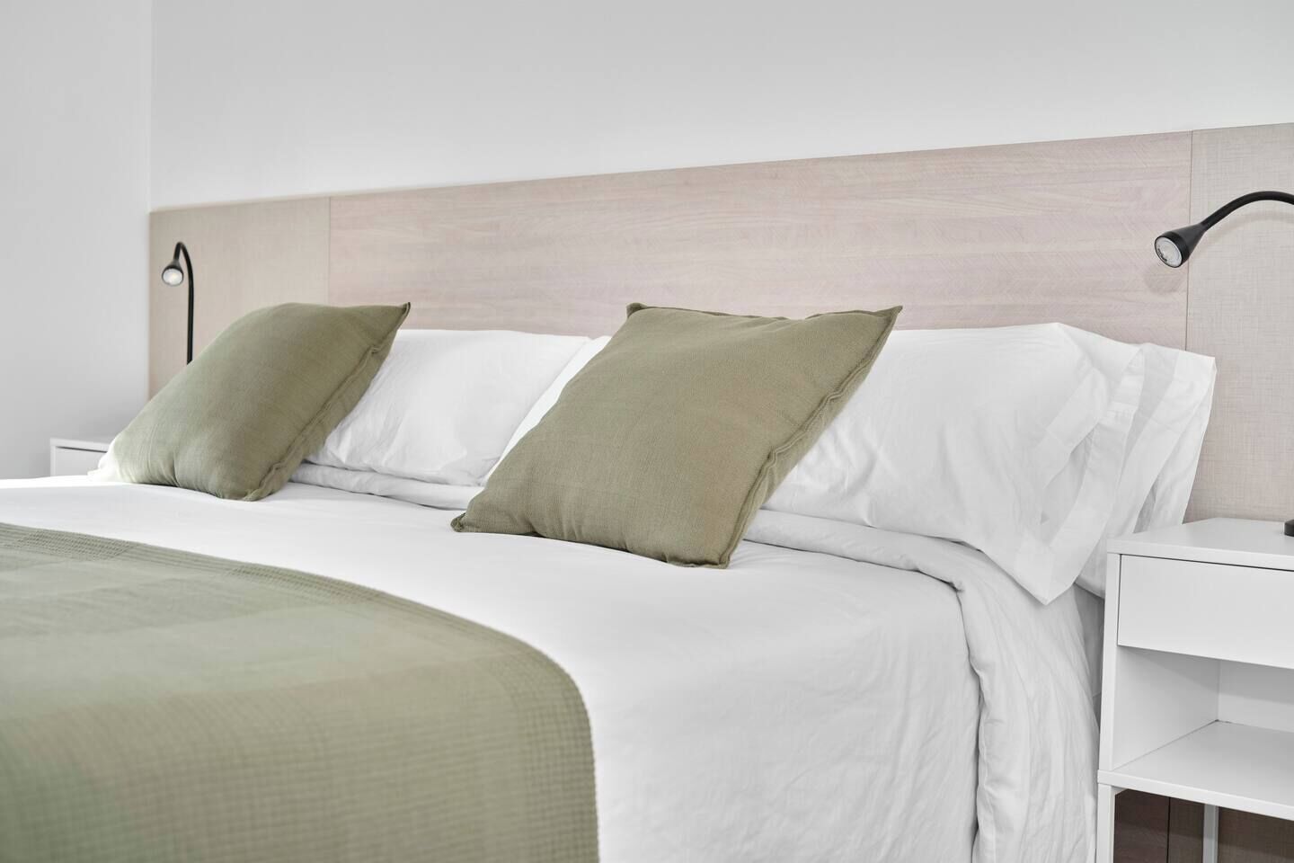 Una cama bien hecha con ropa de cama blanca y verde, una lámpara de noche y una mesita de noche blanca.