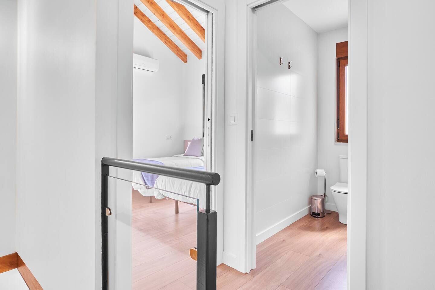 A hallway with a bed in the background, a bathroom with a toilet and a trash bin, and a wooden ceiling beam.