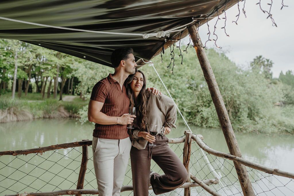 Una pareja está de pie sobre un puente de madera con un dosel de cuerda, con vistas a un río tranquilo y vegetación.
