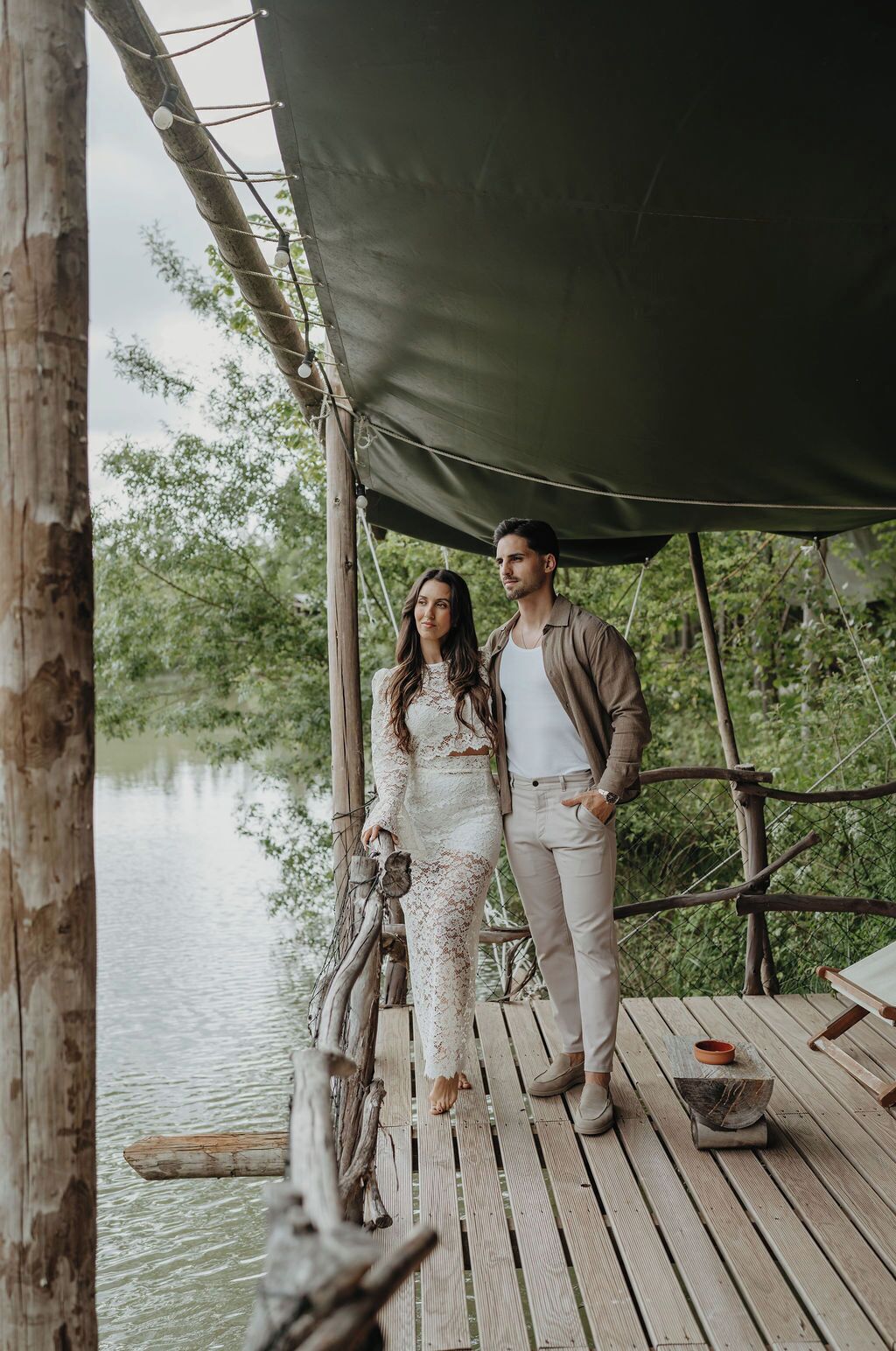 A couple standing on a wooden deck under a canopy, overlooking a body of water.