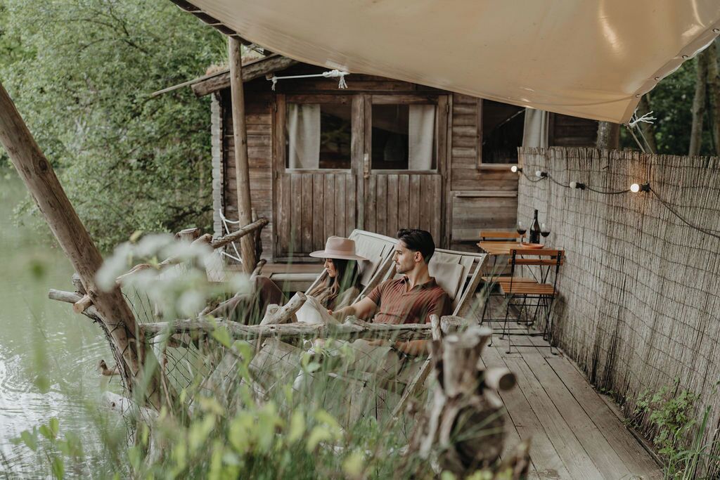 A wooden cabin with a deck overlooking a body of water, featuring a simple outdoor seating area with tables and chairs.