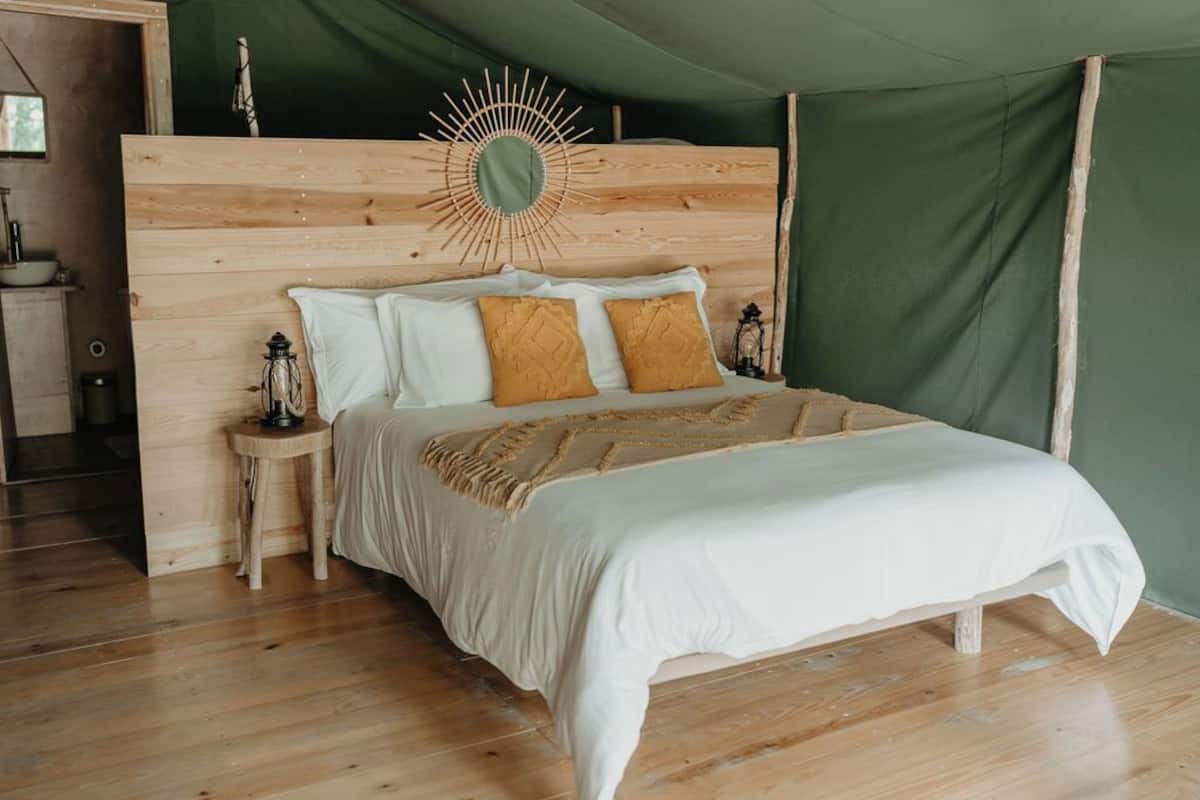 Un lit en bois avec des oreillers blancs et moutarde, une structure verte en forme de tente et une petite table d’appoint avec une lanterne.