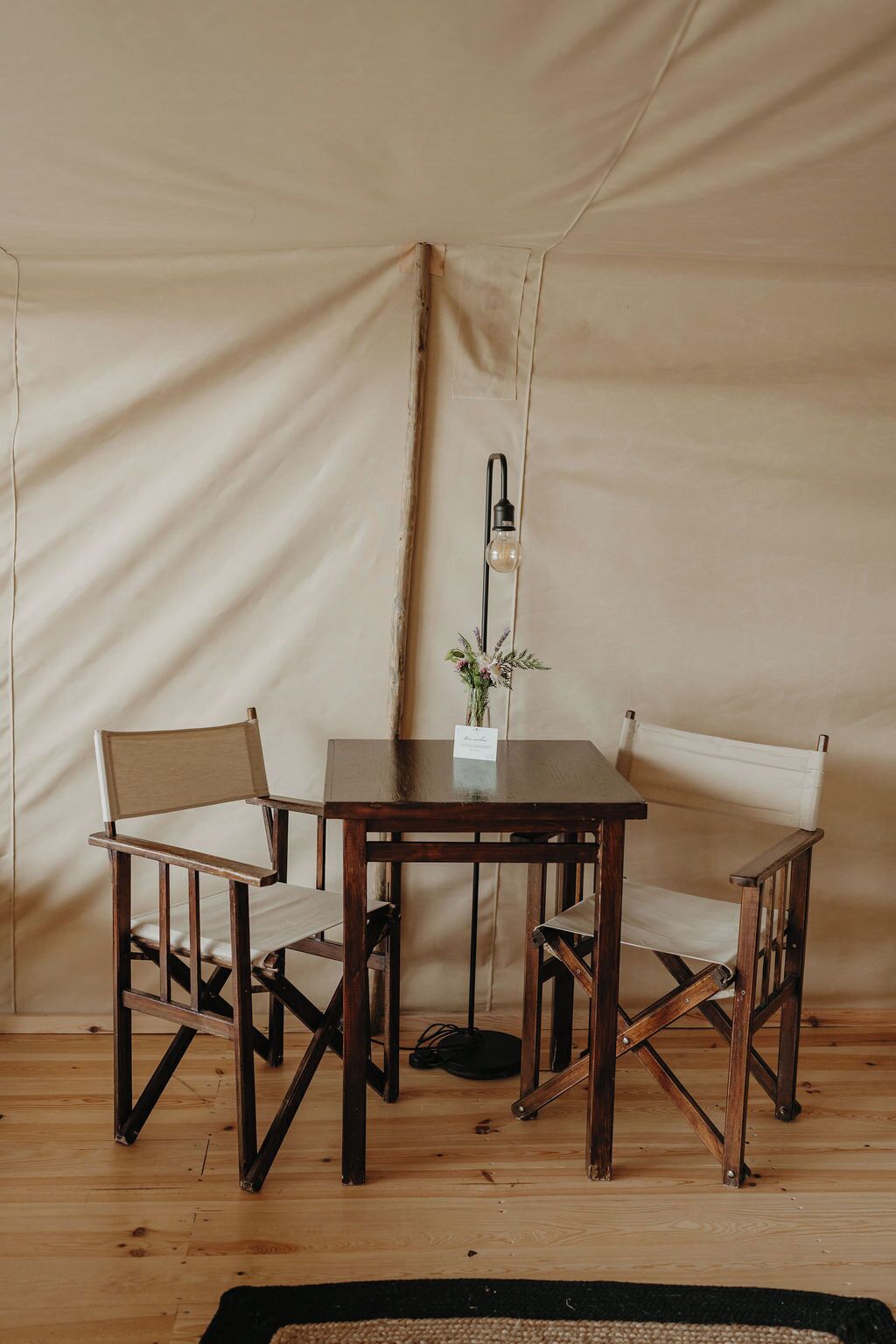 A tent with a wooden table and two chairs, a hanging light fixture, and a small vase with flowers.