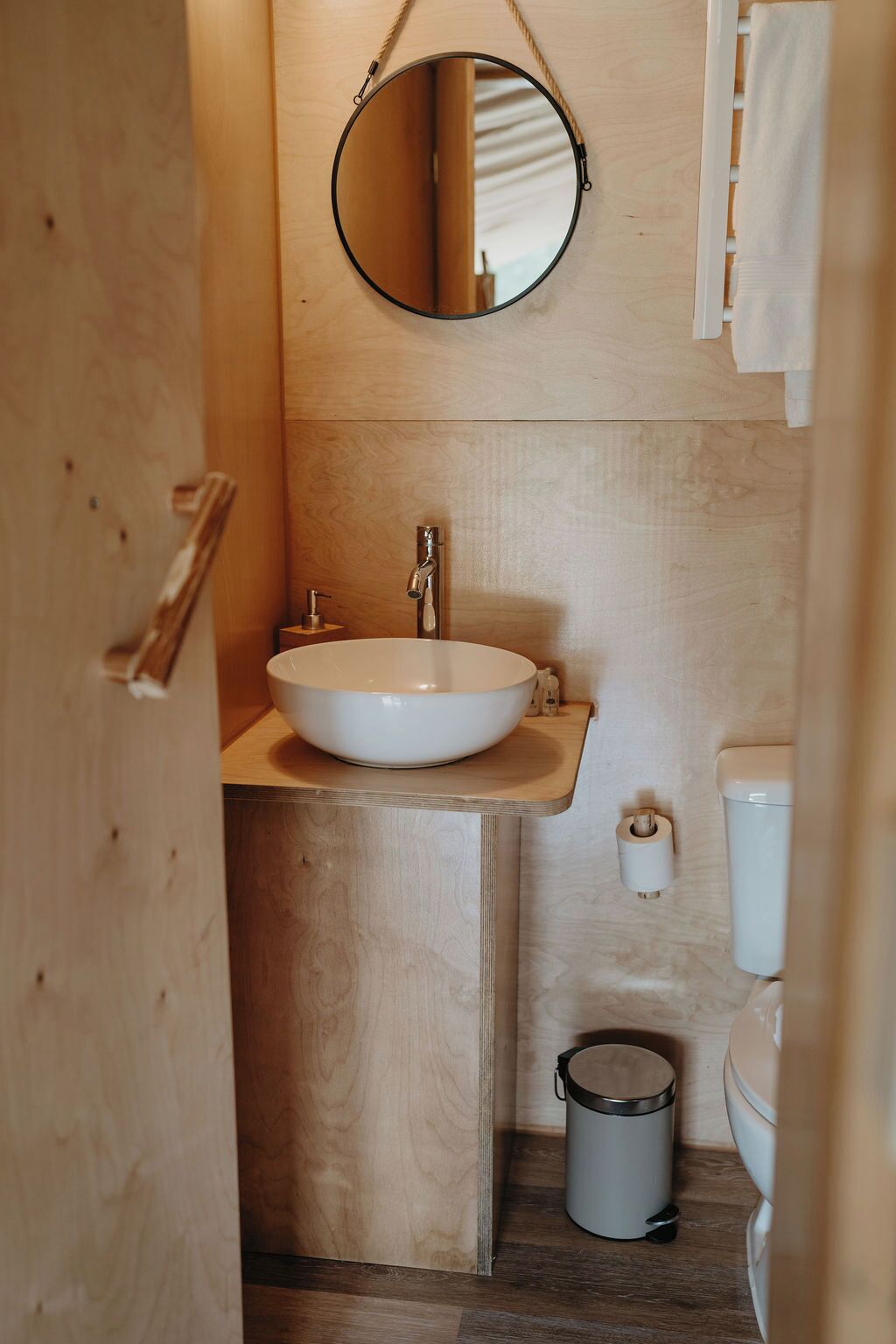 Un baño pequeño con tocador de madera, un espejo redondo, un lavabo blanco, un inodoro y un cubo de basura.