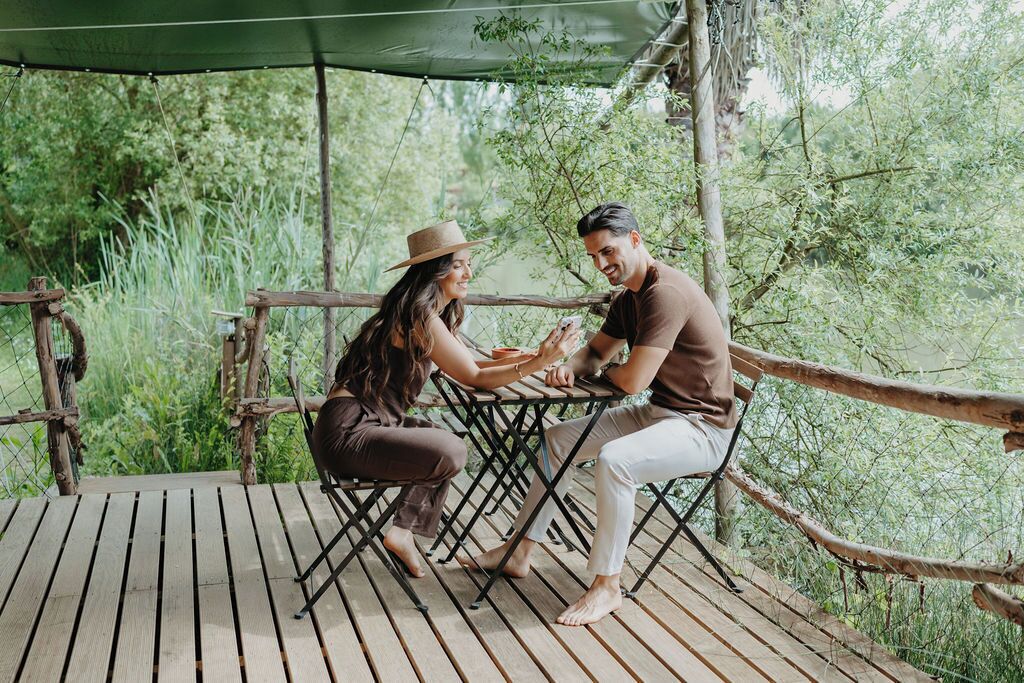 A couple sitting at an outdoor wooden table with a green canopy, surrounded by lush greenery.