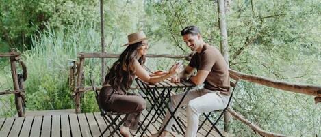 A couple sitting at an outdoor wooden table with a green canopy, surrounded by lush greenery.