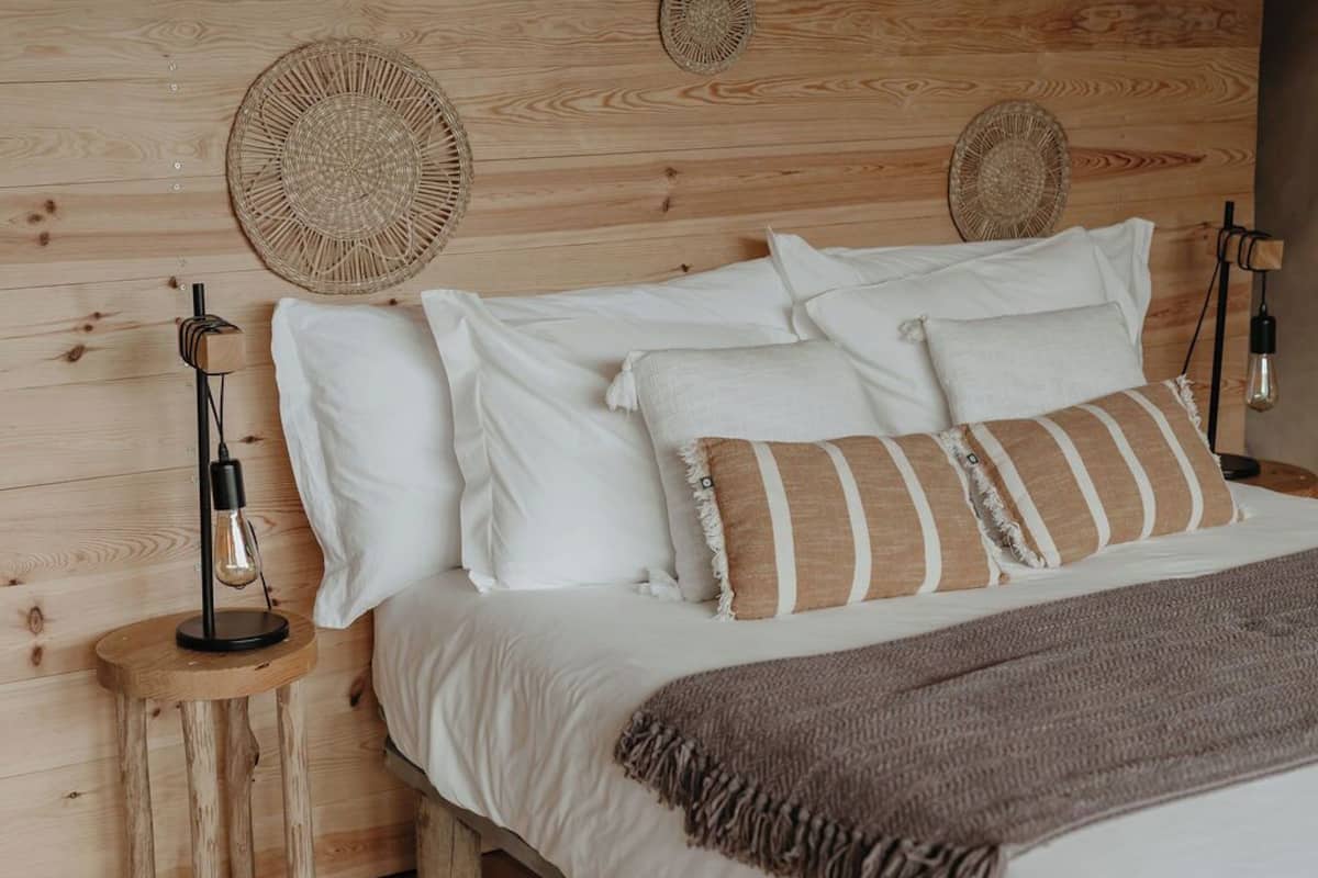 Un lit en bois avec des oreillers blancs et rayés, une tête de lit en osier et une lampe de chevet posée sur une table de nuit en bois.
