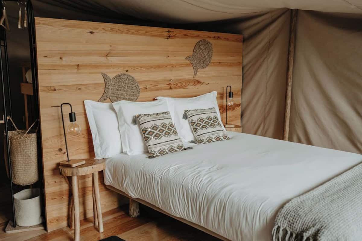 Un lit avec une literie blanche et des oreillers à motifs, une tête de lit en bois agrémentée d’œuvres d’art murales, et une petite table de chevet en bois avec une lampe et un panier en osier.