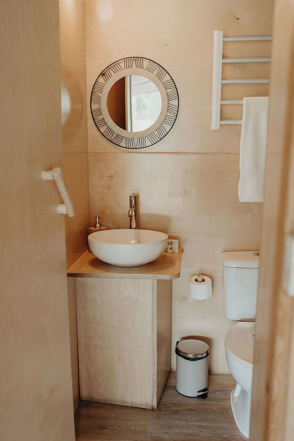 Un baño pequeño con tocador de madera, un espejo redondo, un inodoro y un cubo de basura.