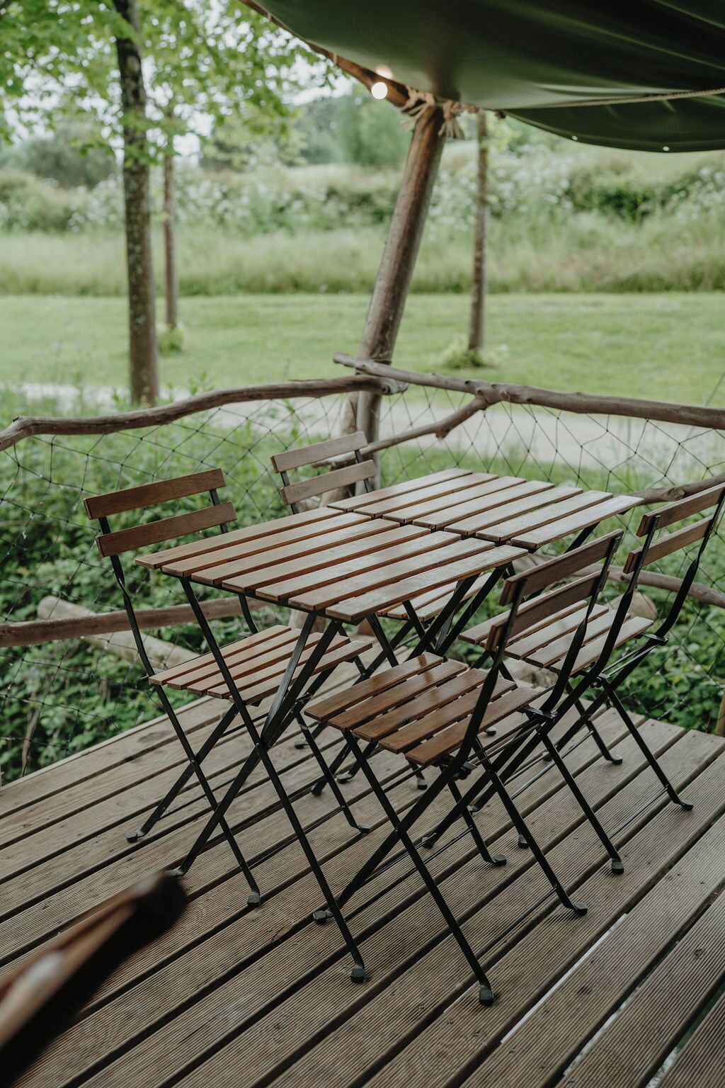 Eine Holzterrasse mit Metallgestellen für Tische und Stühle, umgeben von einem Drahtzaun und Bäumen.