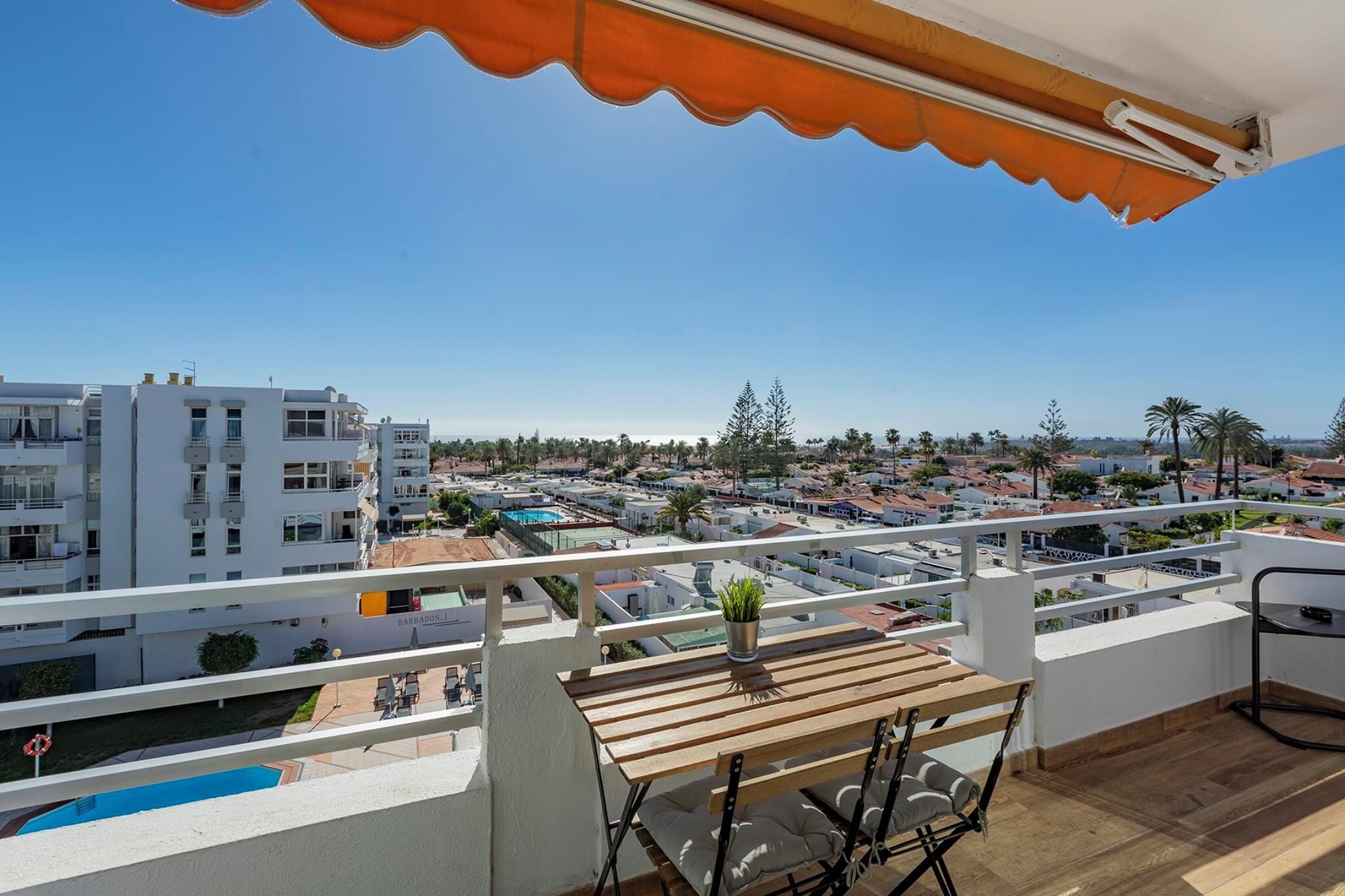 Ein Balkon mit Holztisch, einer Topfpflanze und Blick auf eine Wohngegend mit Gebäuden und einem Schwimmbad.
