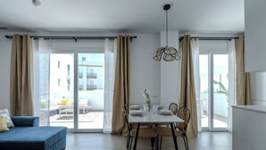 A modern living room with a sofa, a dining table with chairs, and a view through large glass doors to an outdoor balcony.