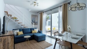 A modern living room with a blue sectional sofa, a dining table with chairs, a staircase with wooden steps, and a large window with curtains.