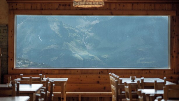 Interior de restaurante con paredes de madera, un ventanal con vistas a la montaña y varias mesas y sillas de madera.