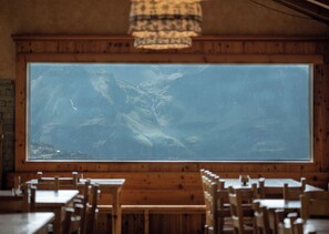 Interior de restaurante con paredes de madera, un ventanal con vistas a la montaña y varias mesas y sillas de madera.