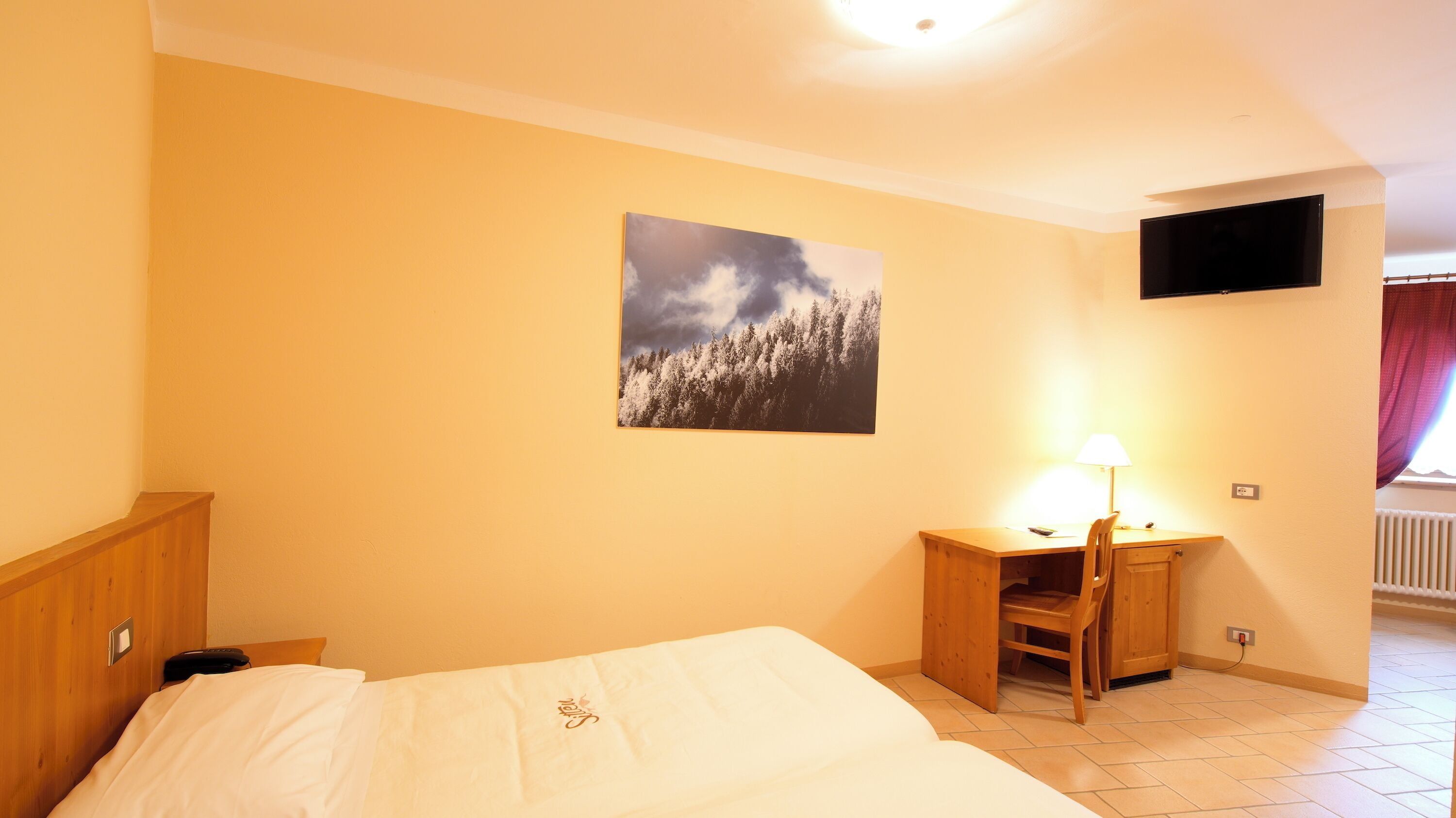 A hotel room with a bed, a desk, a chair, a TV mounted on the wall, and a window with curtains.