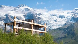 Una cabaña de madera con estructura de techo sencilla, rodeada de vegetación exuberante y montañas nevadas al fondo bajo un cielo azul despejado.