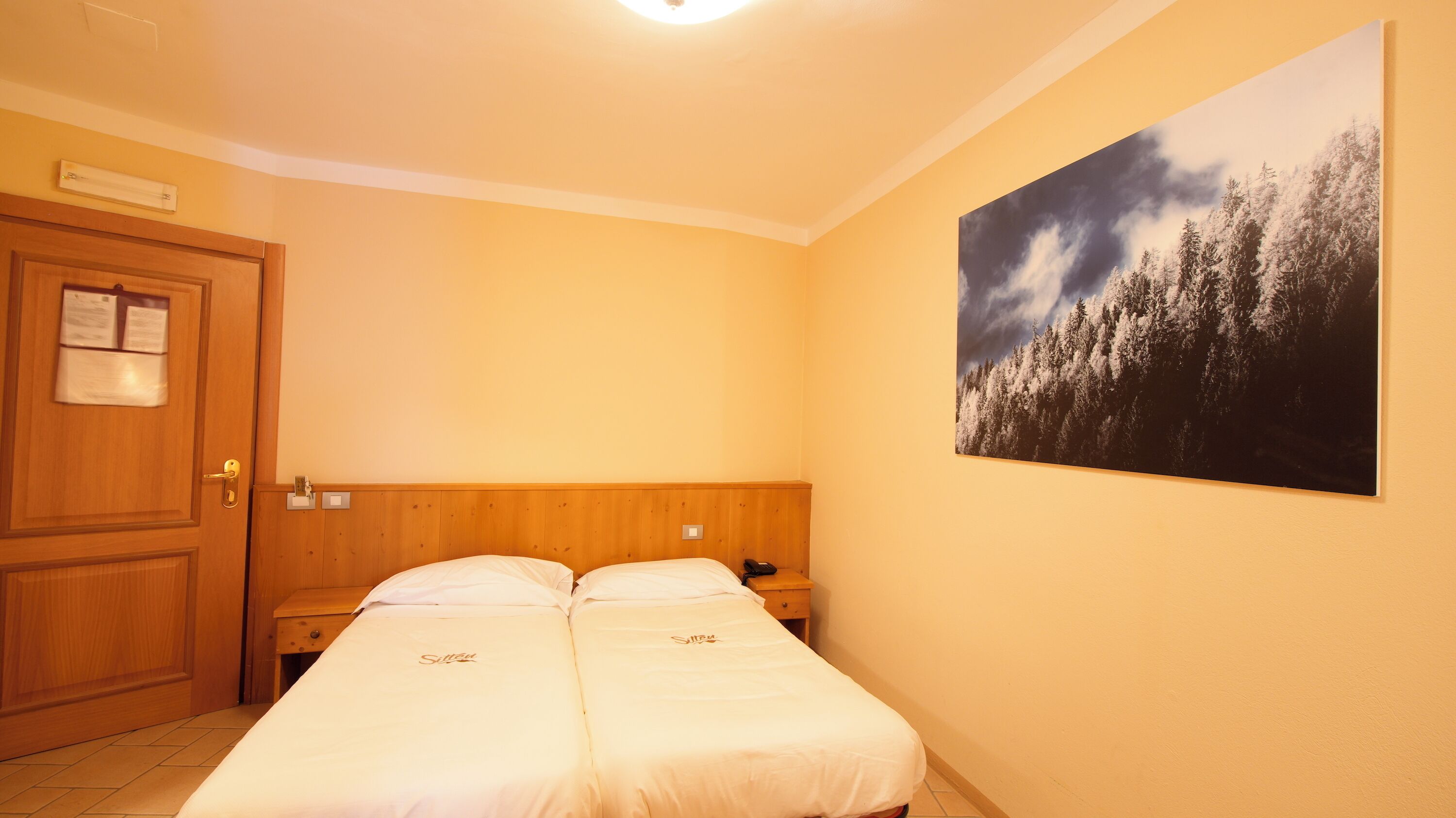 A hotel room with two single beds, a wooden headboard, a wooden door, a wall-mounted light, and a forest landscape painting.