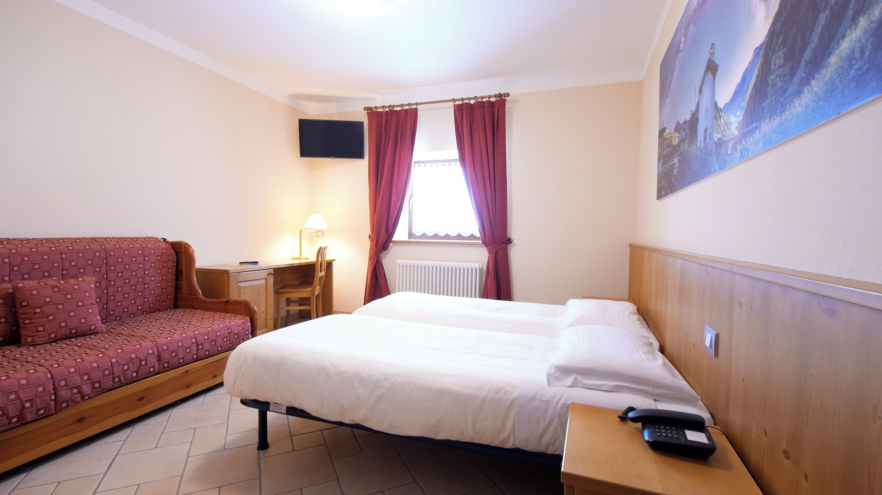 A hotel room with a bed, a desk with a chair, a sofa, a painting on the wall, and a window with curtains.