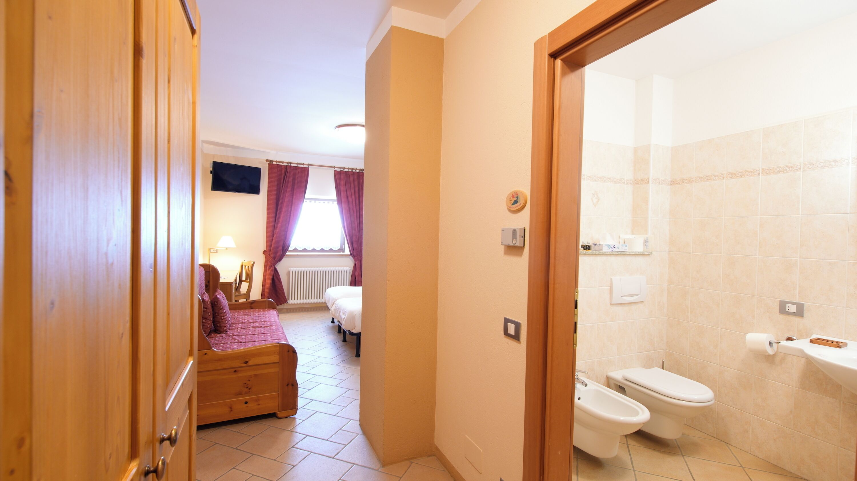A hotel room with a bed, a desk, a chair, a window with curtains, a radiator, a TV mounted on the wall, and a bathroom with a toilet and bidet.