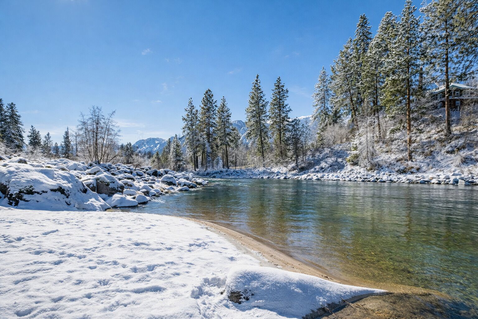A snow-covered riverbank with clear water, surrounded by snow-laden trees and a mountain in the background.